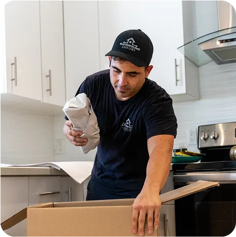 A man wearing a black shirt and cap is opening a cardboard box on a table.